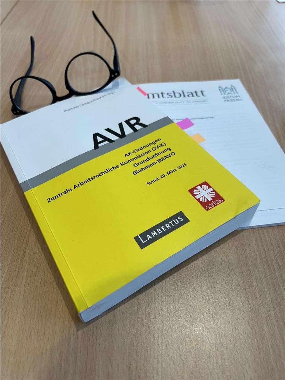 Schwarze runde Brille liegt neben einem gelben Fachbuch mit der Aufschrift „AVR“ auf einem Holztisch. Unter dem Buch liegen markierte Dokumente und Unterlagen zum Arbeitsrecht.